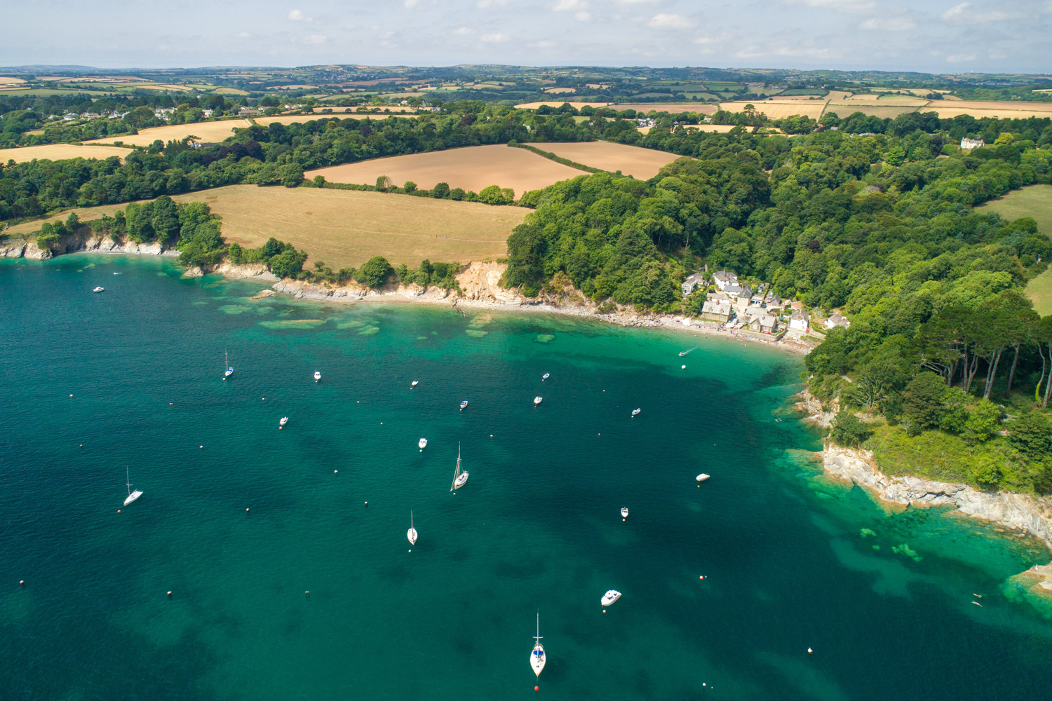 An aerial image of Durgan on the Helford River a small hamlet of buildings seen from above with blue water and a small sandy beach. There are boats on the water